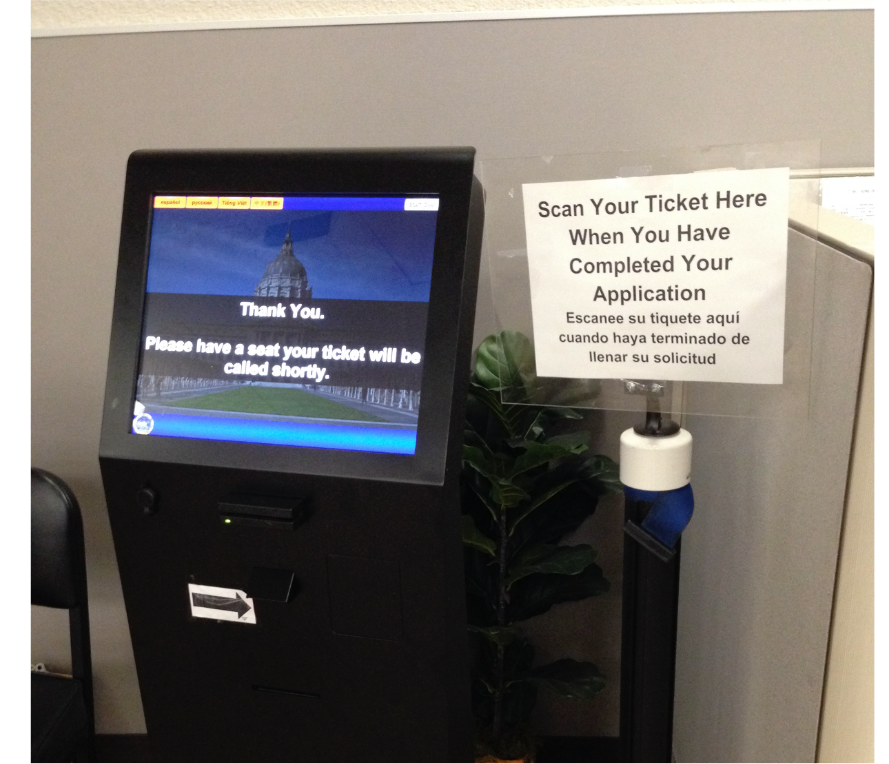 Photo of a black, digital kiosk on the left with words on the touch screen reading, "Thank you. Please have a seat your ticket will be called shortly." A paper sign on a pole next to it says, "Scan Your Ticket Hear When You Have Completed Your Application." A Spanish translations of these words are beneath it. 